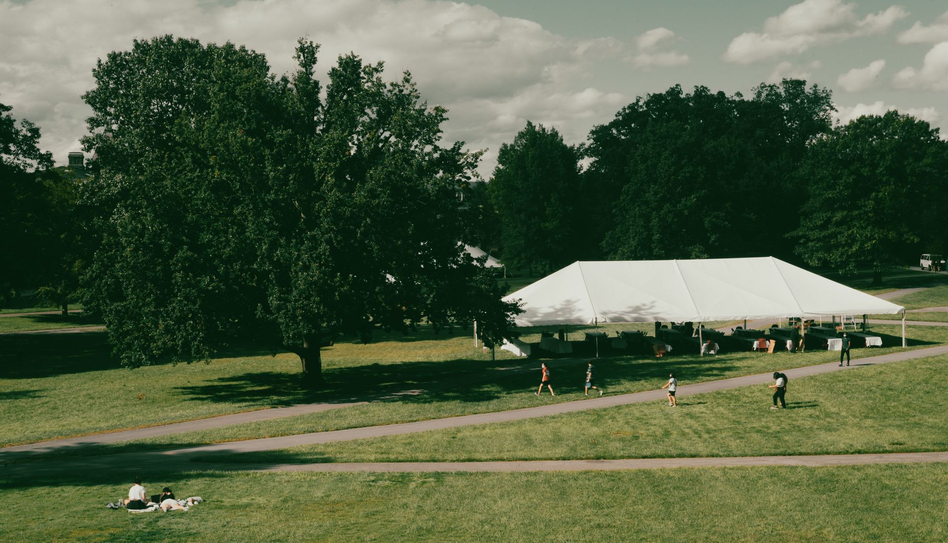 Aerial photo of a park with a large white tent, people enjoying greenery and blue skies.